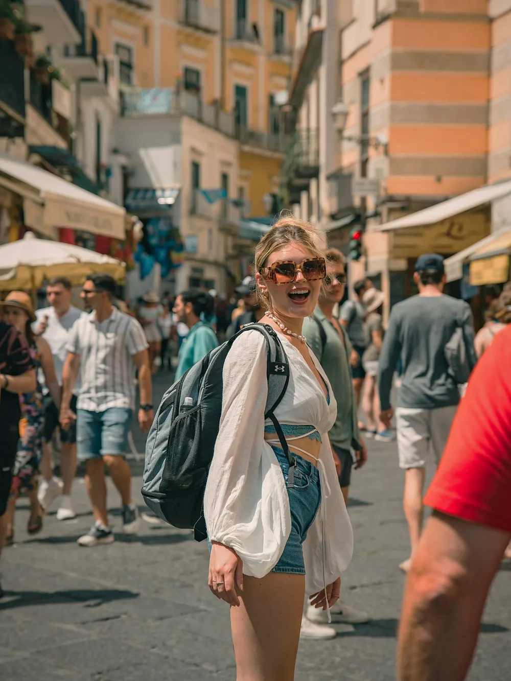 A woman in white smiling on a busy Amalfi street with passers-by behind her